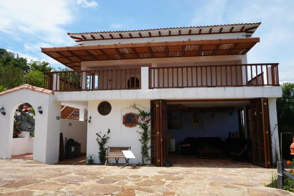 Fachada blanca de la casa de dos pisos, con balcón de madera y patio de piedra