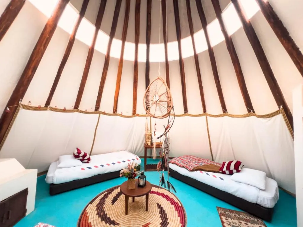 Interior del tipi: camas tendidas, piso turquesa, tapete tejido y la estructura de madera que sube hacia la claraboya