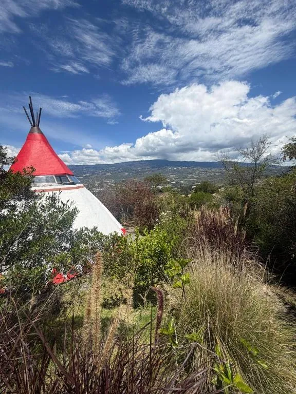 Tipi de techo rojo entre la vegetación, con el valle de Villa de Leyva y el cielo de fondo